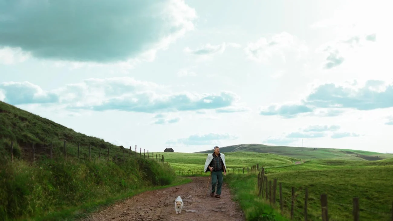 A man walking on a dirt path with a small dog in a lush green countryside under a partly cloudy sky.