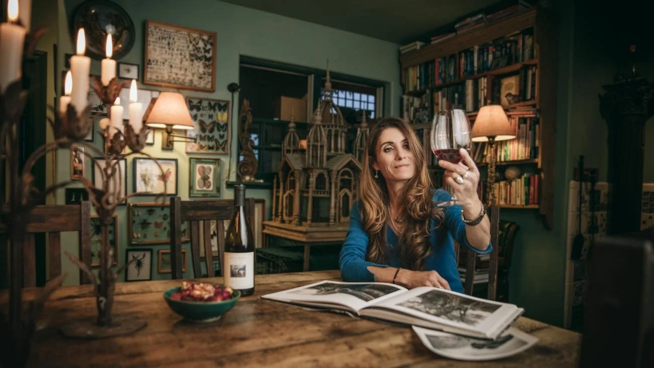 Woman sitting at a wooden table in a cozy, art-filled room, holding a glass of red wine, with an open book and a bottle of wine in front of her, surrounded by books and artwork.