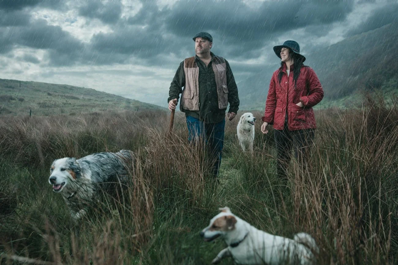 Two people, a man and a woman, walking through tall grass during a rainstorm with three dogs in a moody, cloudy outdoor landscape.