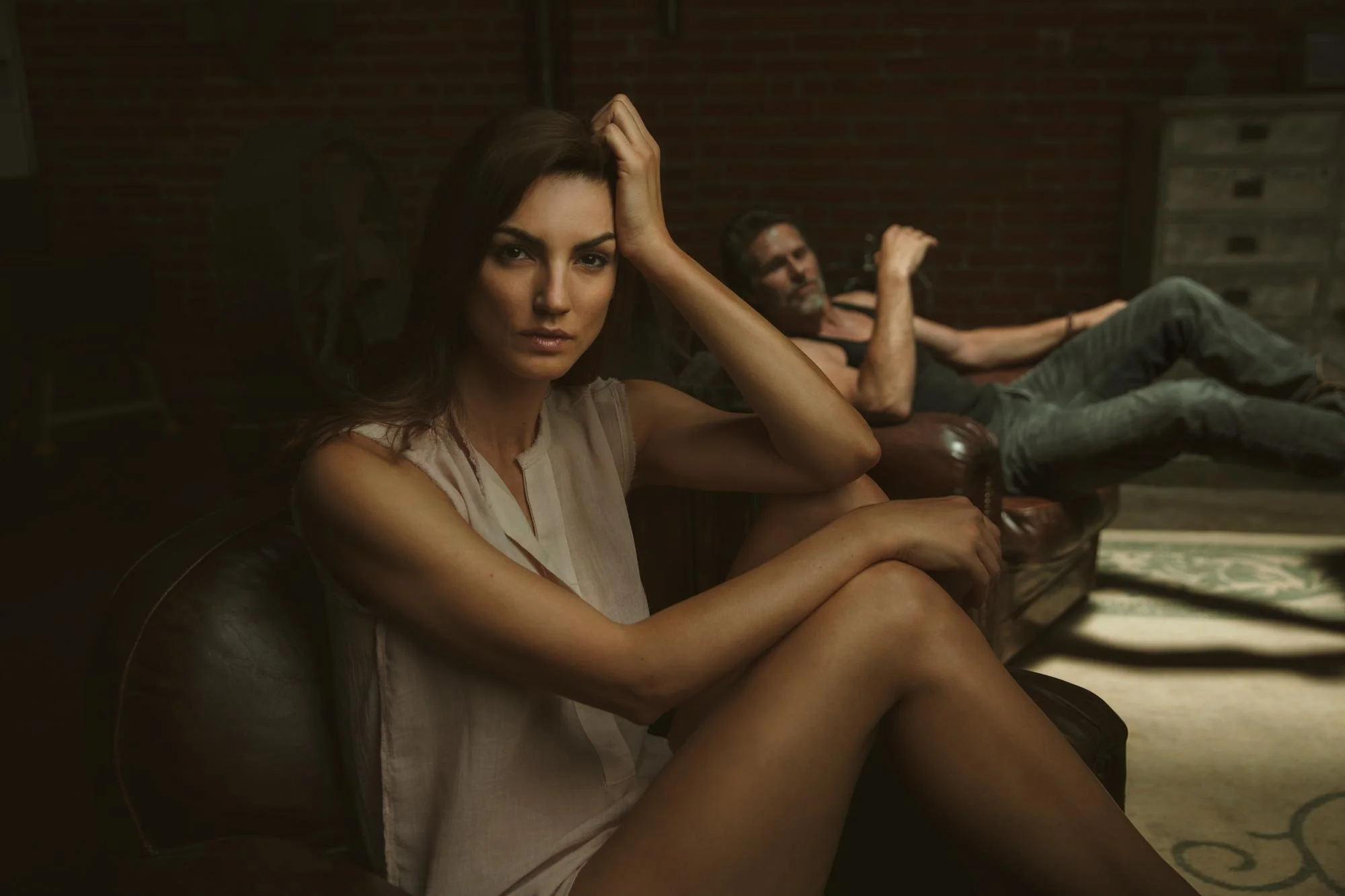 A woman with dark hair sitting on a leather chair in the foreground, looking at the camera with a thoughtful expression, while a man with gray hair and a beard lounges on a couch in the background in a dimly lit room with brick walls.