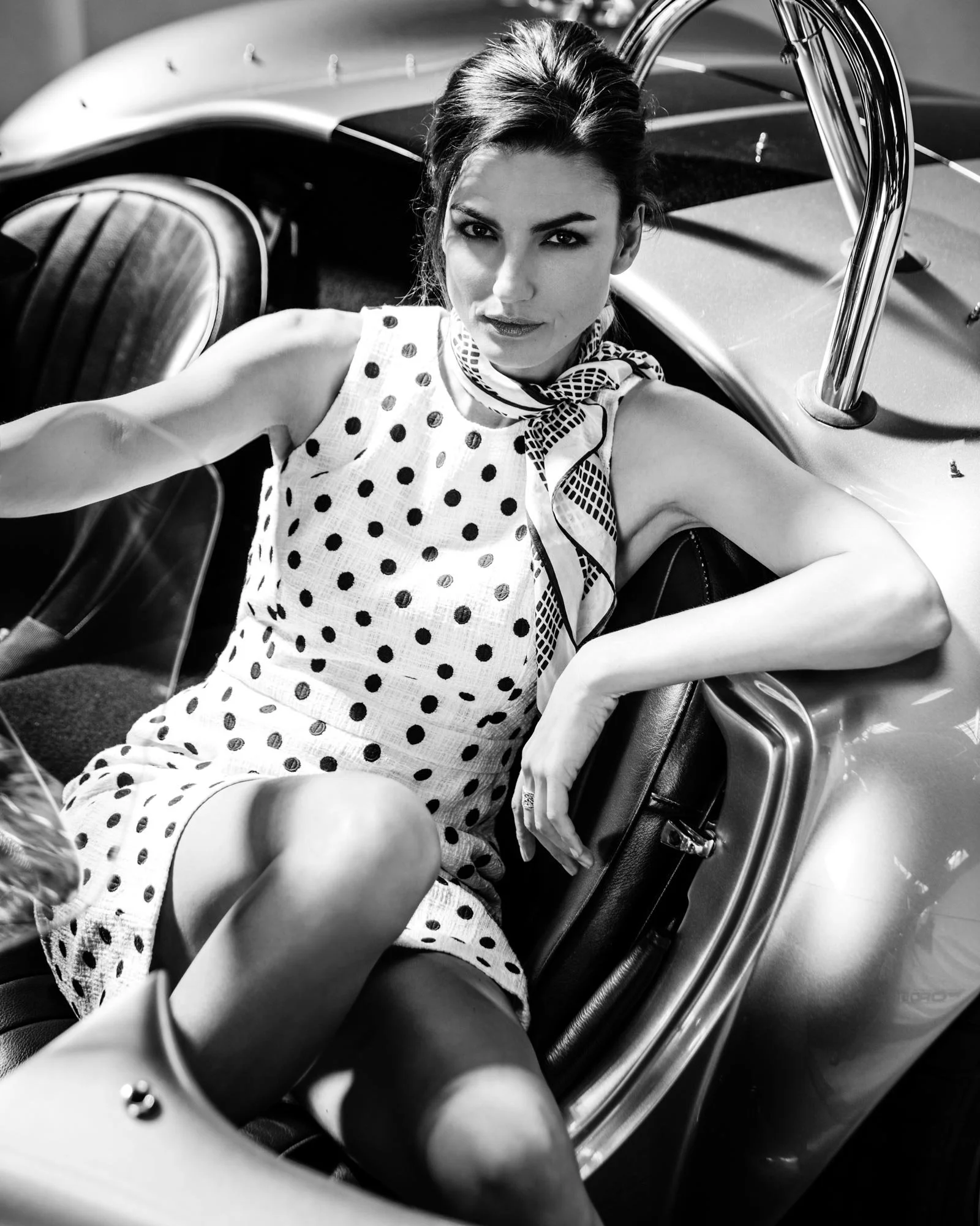 A woman in a polka dot dress and a scarf around her neck, sitting in a classic convertible car with her arm resting on the door.