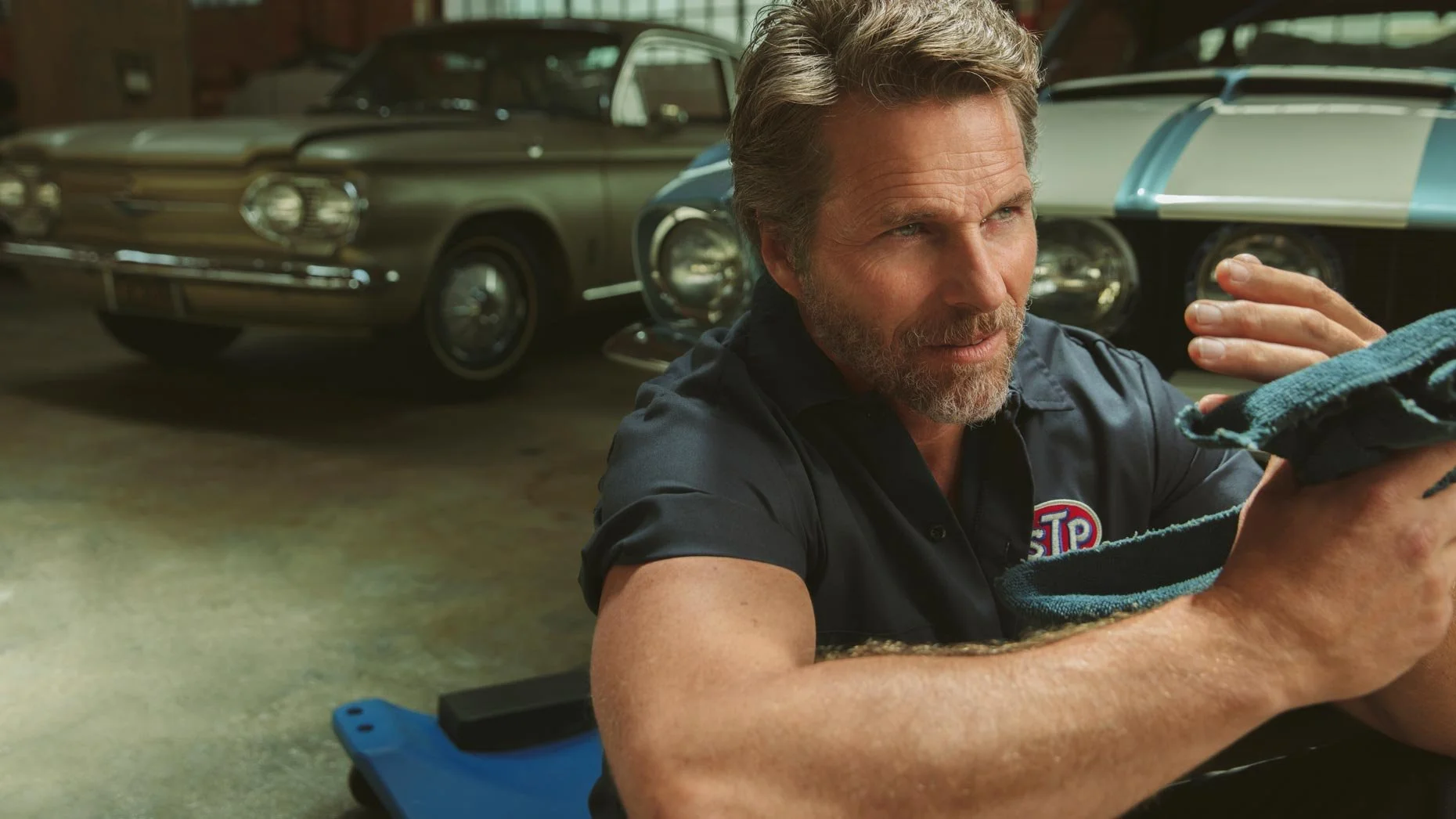 A man with a beard and short hair cleaning a car part in a garage, with classic cars parked in the background.