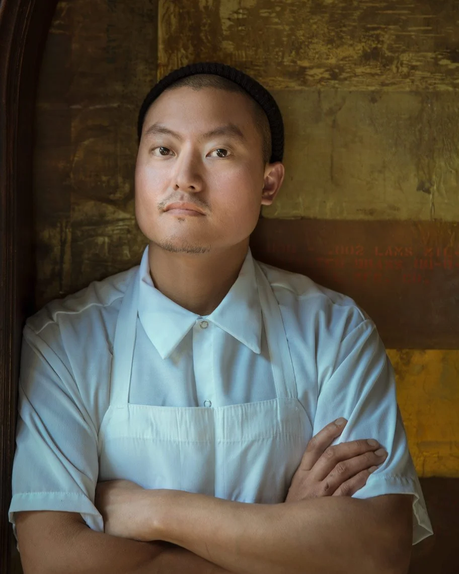 A man in a white chef's coat and apron crosses his arms and looks directly at the camera, standing against a rustic wooden background.