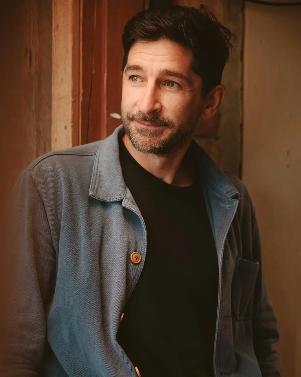 Portrait of a man with dark hair and a beard, wearing a gray jacket over a black shirt, looking to the side with a slight smile, standing in front of a wooden wall.