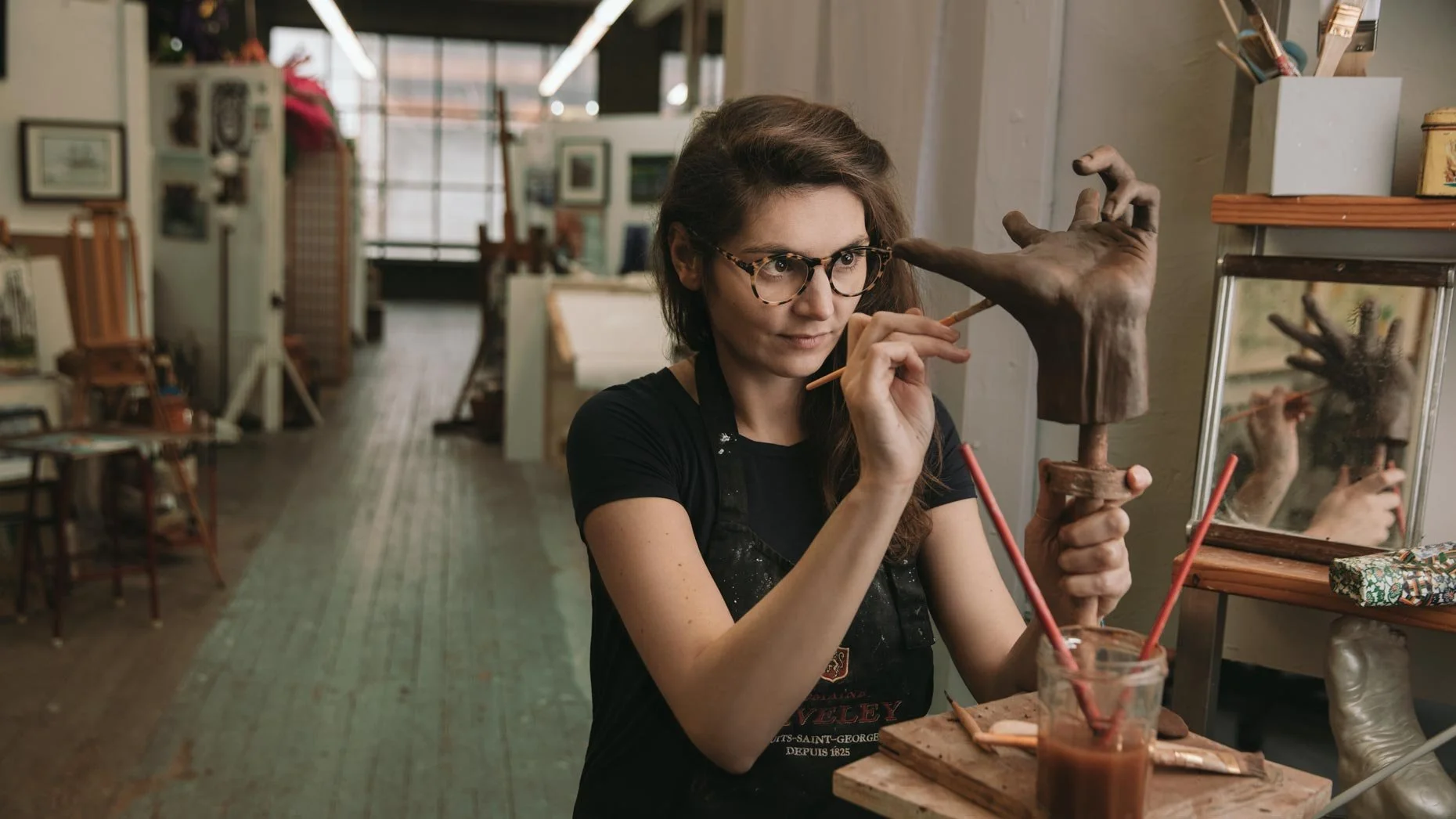 A woman with glasses and dark hair working on a sculpture of a hand in an art studio.