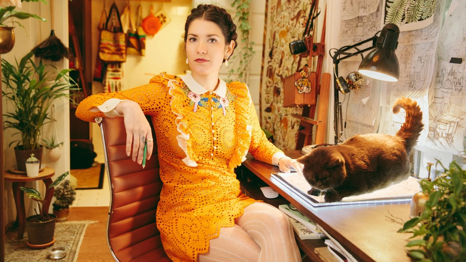 A woman with braided hair wearing an orange lace dress sitting at a desk with a black cat lying on a drawing table. The room has plants, artwork, and a lamp.