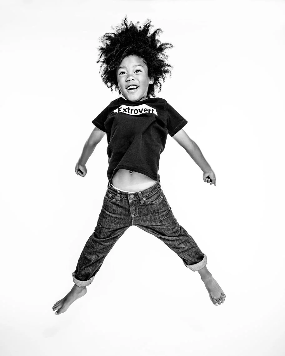 A young girl with curly hair jumping joyfully with arms and legs spread wide, wearing a t-shirt with the word 'Extrovert' on it, and jeans, against a plain background.