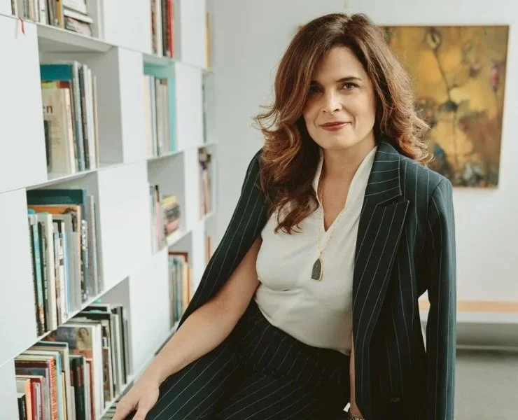 A woman with wavy brown hair and light skin, wearing a pinstripe suit over a white blouse with a pendant necklace, sitting next to a bookshelf filled with books in a library or office setting.