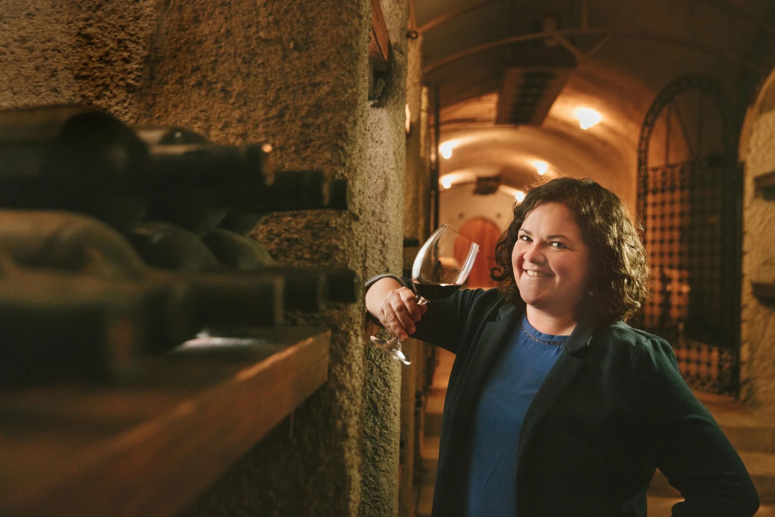 Lifestyle portrait of a smiling woman holding a glass of red wine in a stone-arched wine cave, Napa Valley – Marc Olivier Le Blanc Photography