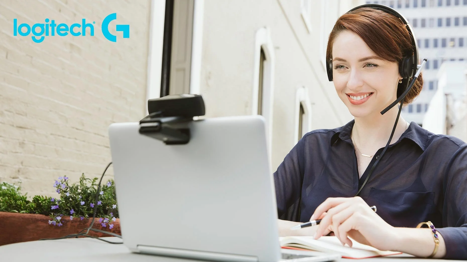 Advertising photo of a smiling woman using a Logitech webcam and headset at a laptop outdoors – Marc Olivier Le Blanc Photography