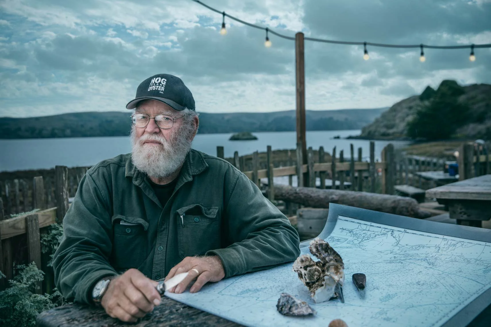 An older man with glasses and a beard wearing a cap and jacket sitting at a table with bones, rocks, and a detailed map outdoors near a body of water with hills in the background, under string lights.