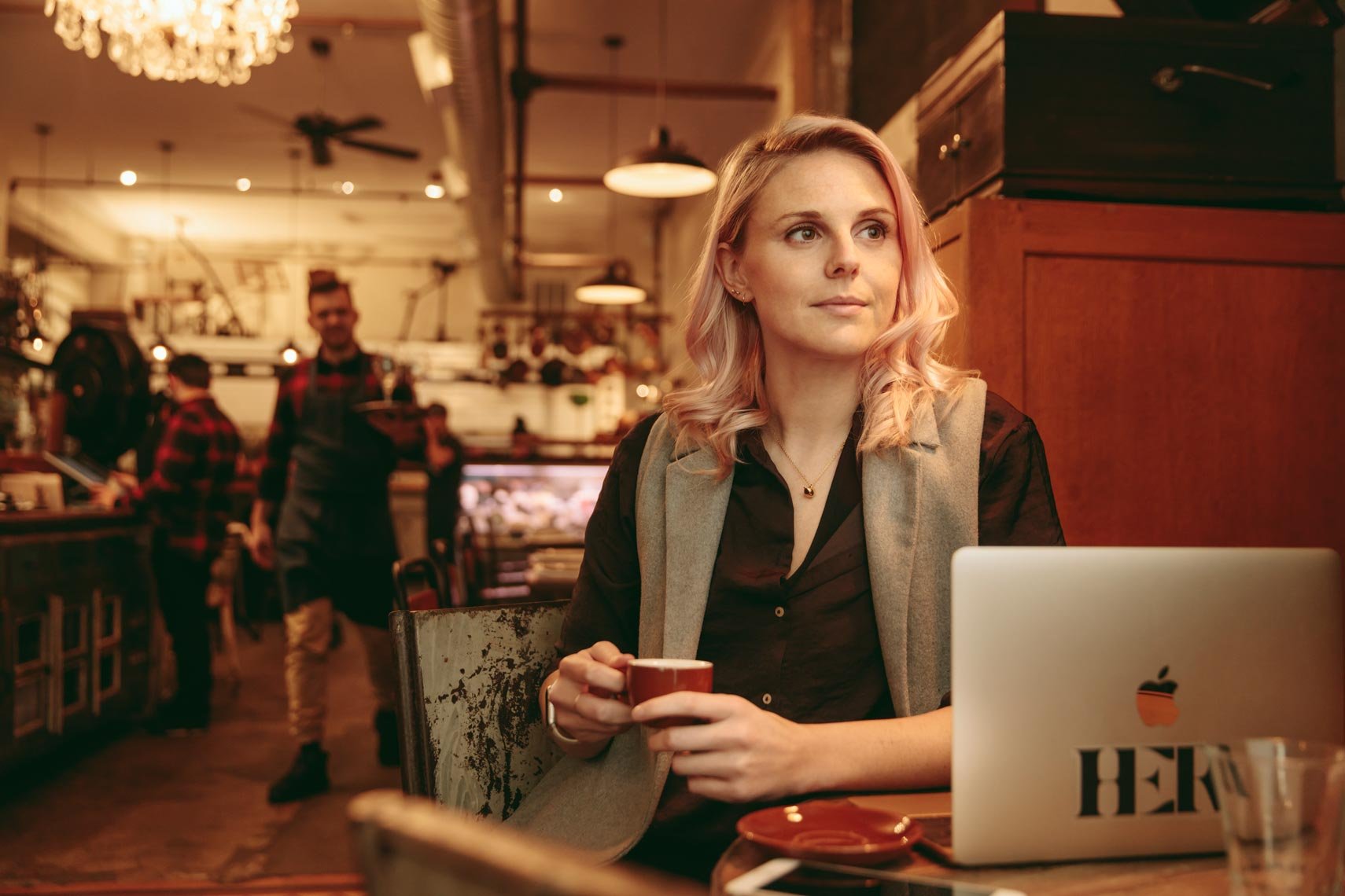 A woman with blonde hair sitting at a coffee shop table, holding a cup, with a laptop in front of her, and a background of staff working behind the counter.