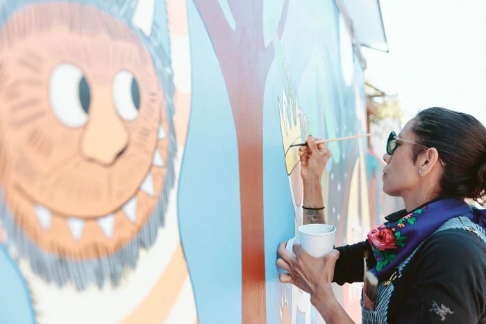 A woman painting a colorful mural on a wall, holding a small bowl and using a thin paintbrush, with her hair in a ponytail and wearing glasses and a scarf.
