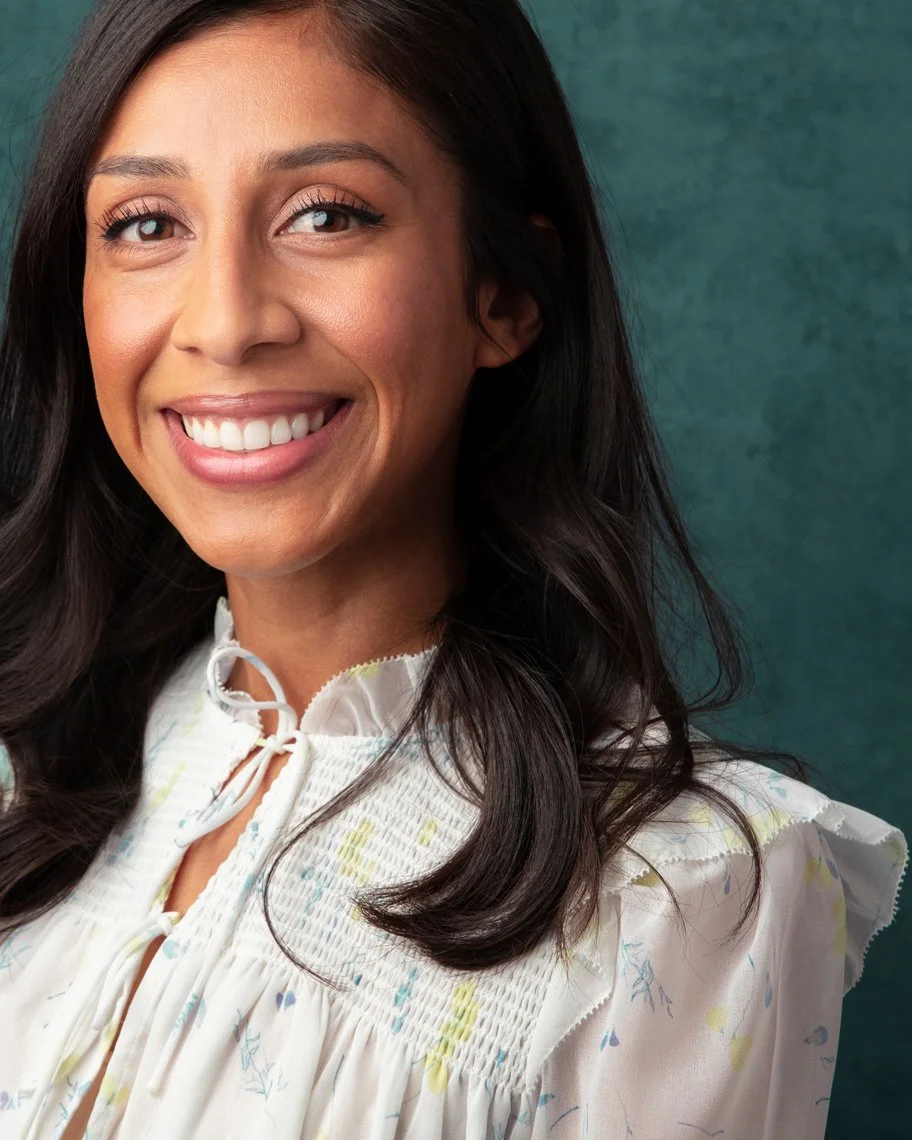A woman with long dark hair smiling, wearing a white blouse, against a green background.
