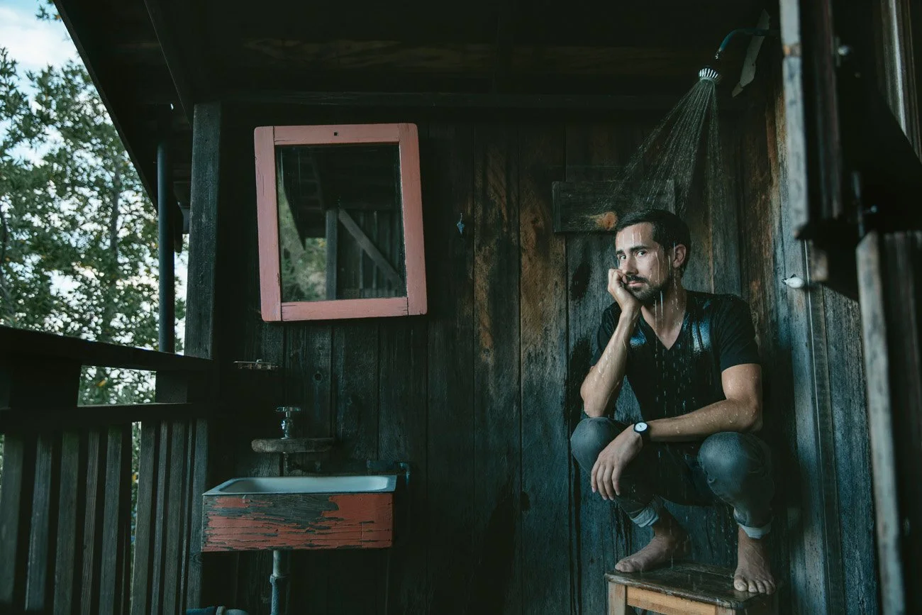 Moody editorial portrait of a man sitting under an outdoor shower at a rustic cabin – Marc Olivier Le Blanc Photography