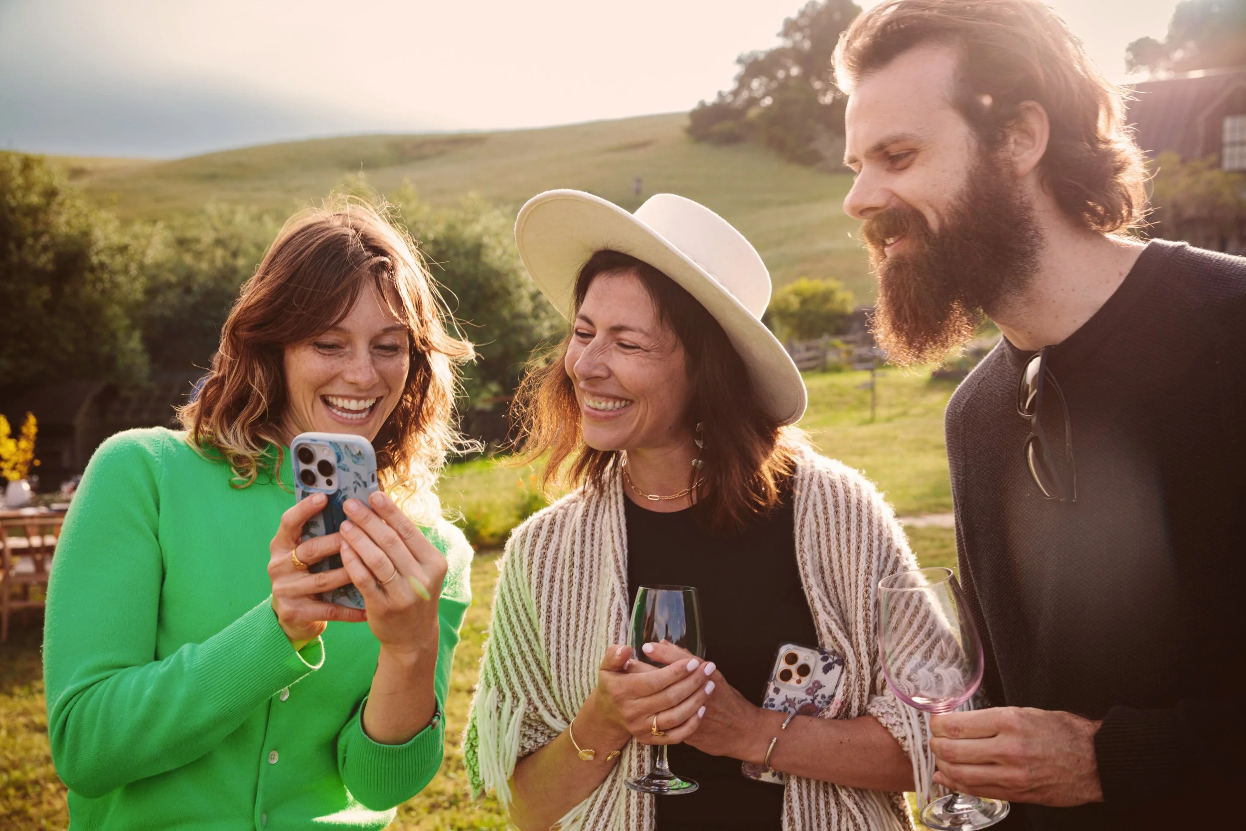 Lifestyle photo of three friends laughing together with wine glasses at golden hour in a rolling green Bay Area landscape – Marc Olivier Le Blanc Photography