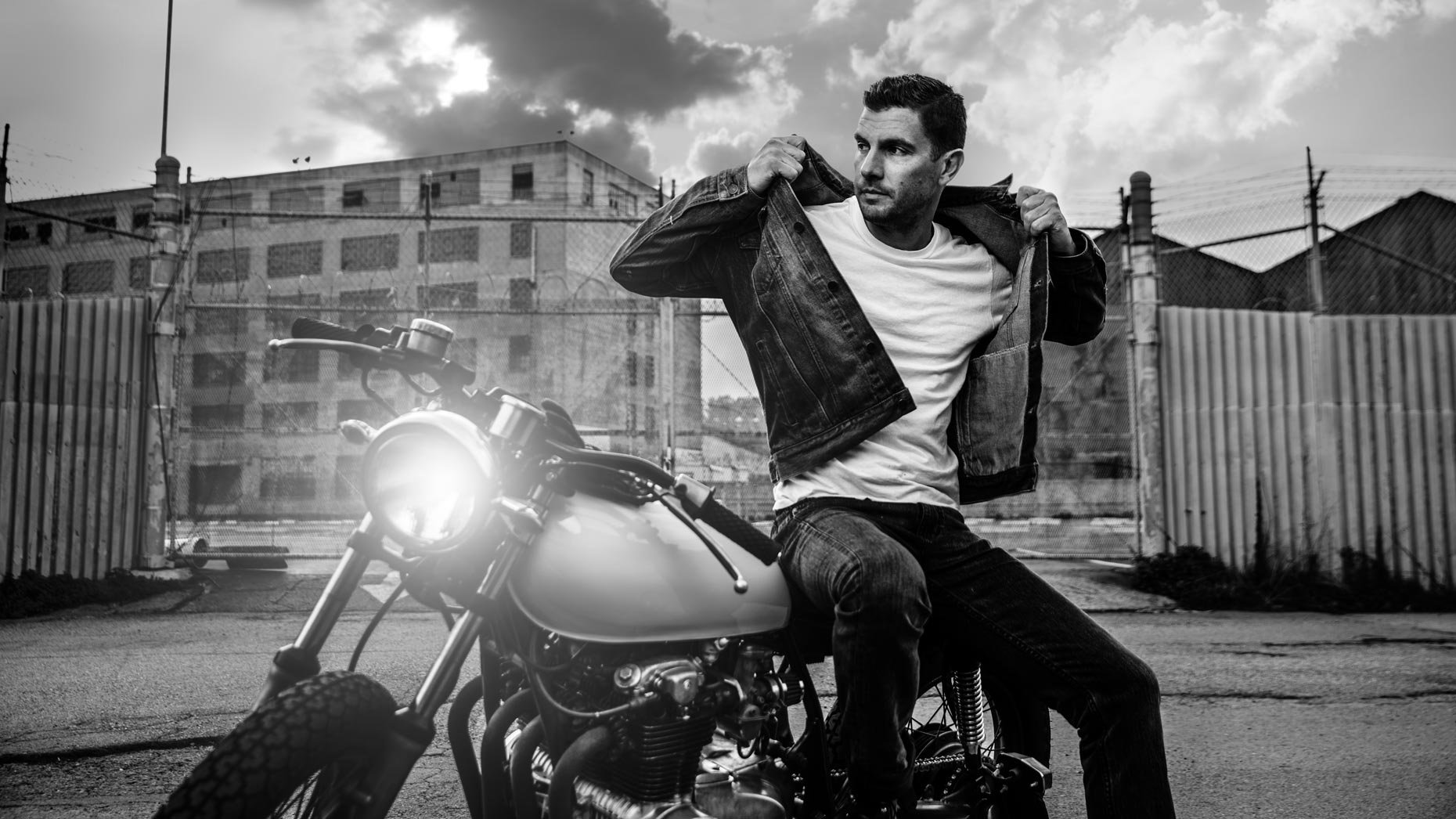 A man adjusting his denim jacket while sitting on a motorcycle in an urban setting with a cloudy sky.