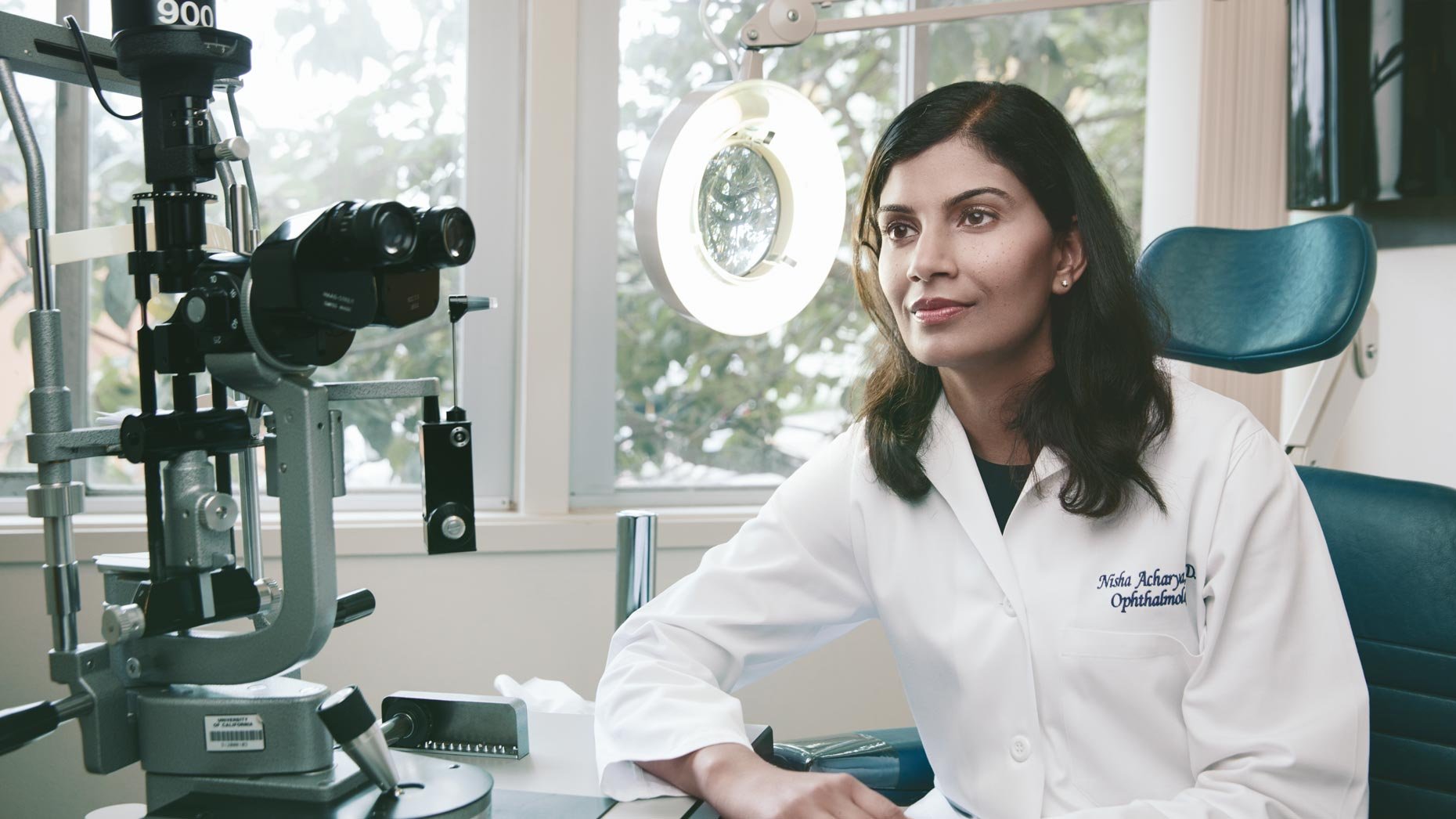 Ophthalmologist sitting at her desk in an eye clinic, with examination equipment and a bright ring light behind her, looking thoughtfully to her right.
