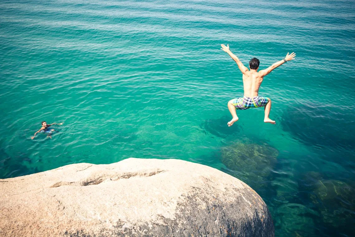 A man jumping off a rock into clear water while another person swims nearby.