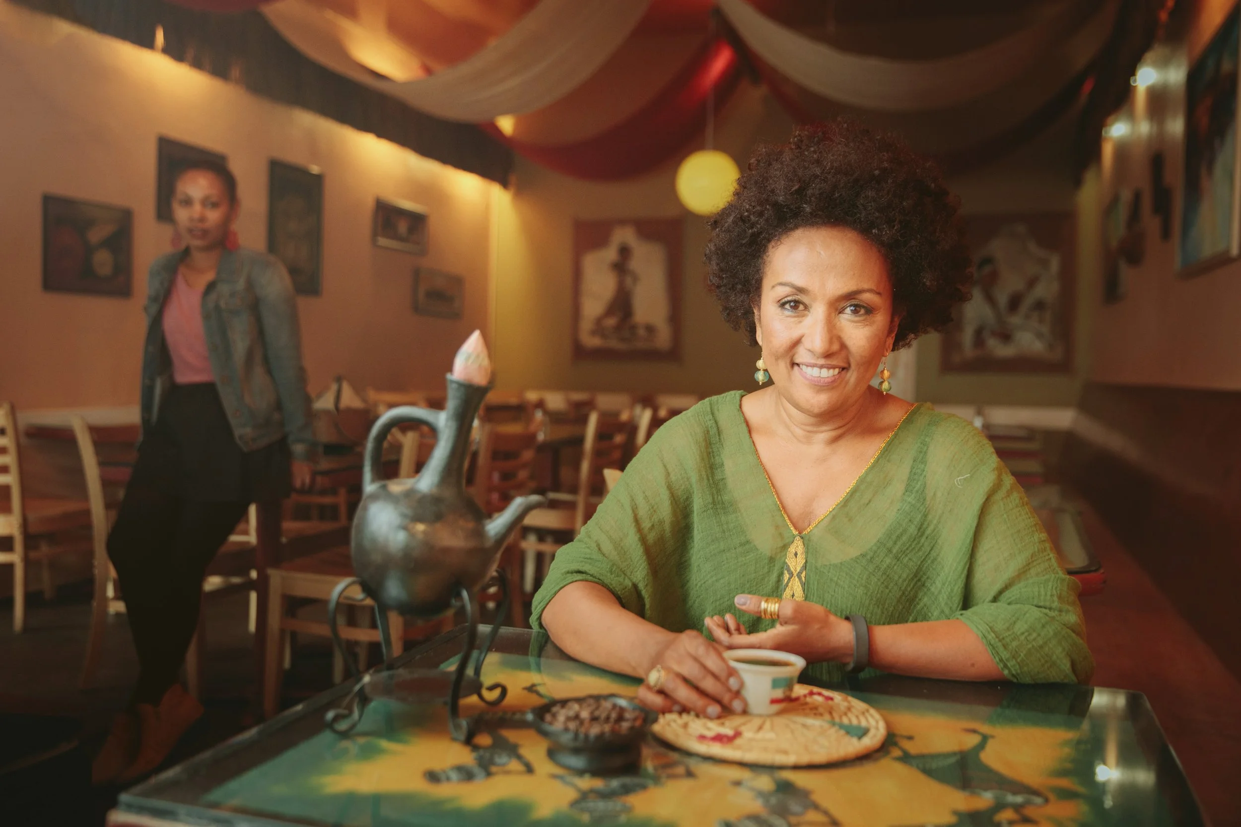 Editorial portrait of a smiling woman holding a coffee cup in a warmly lit San Francisco Ethiopian restaurant – Marc Olivier Le Blanc Photography