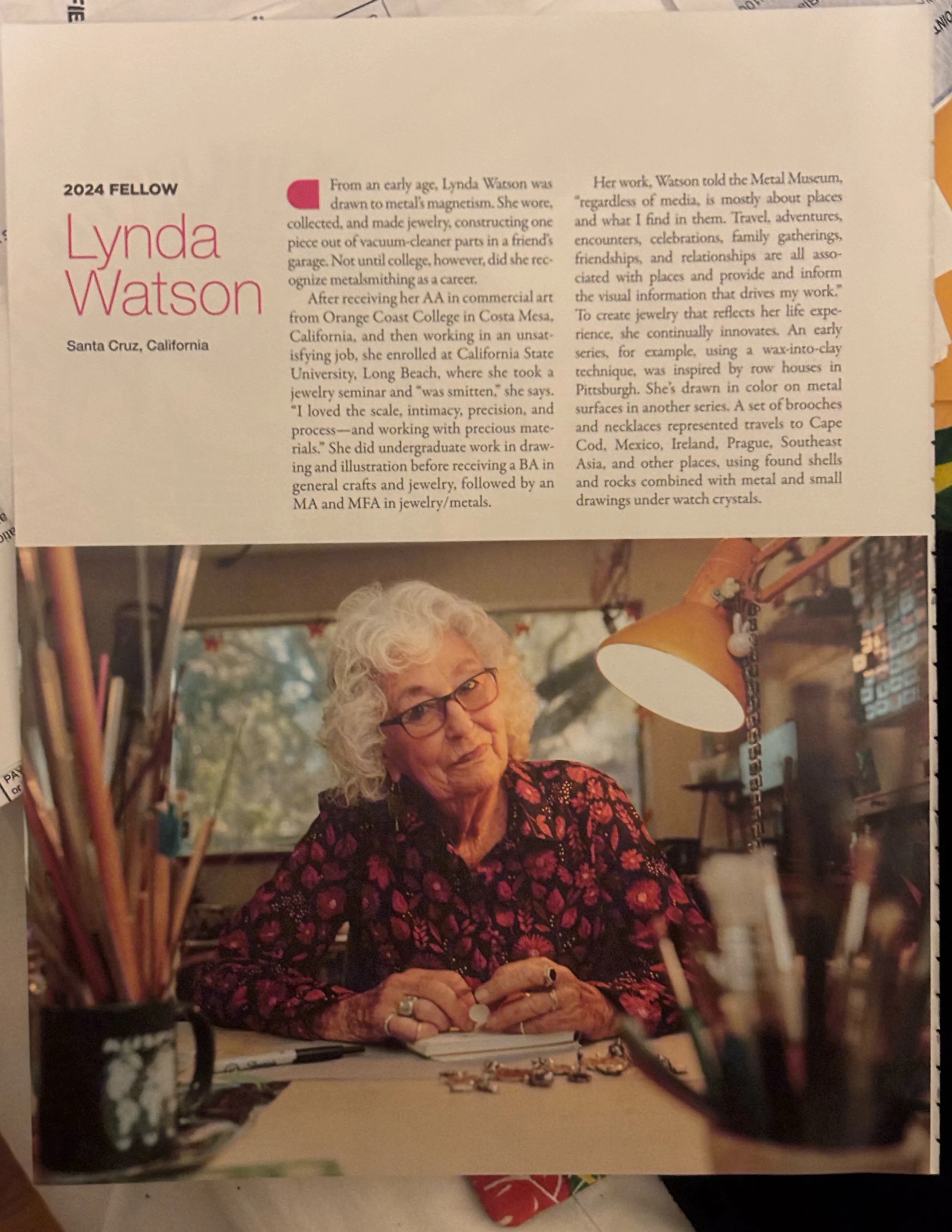 A woman with curly gray hair, glasses, and wearing a black dress with a red floral pattern, sitting at a table in a room with bookshelves and decorations. She is looking at jewelry on the table and holding a jewelry piece.