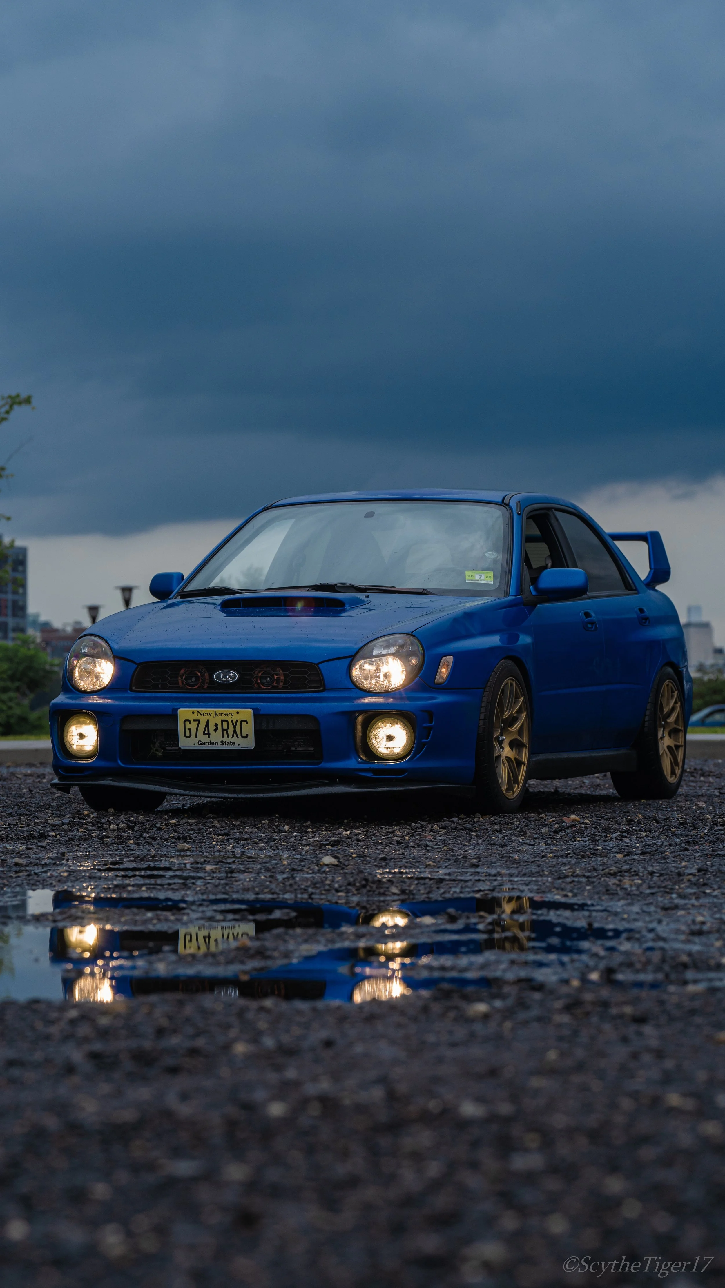 A blue Subaru WRX car parked on a gravel surface after rain, with a puddle reflecting the front of the car. The sky in the background is cloudy and dark.