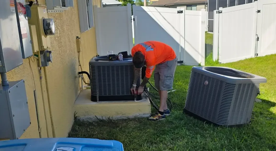 An Affordable Air HVAC technician in a high-visibility orange company shirt works on an outdoor AC condenser. A new unit sits nearby on the grass, ready for installation outside a home with a yellow exterior and white fence.