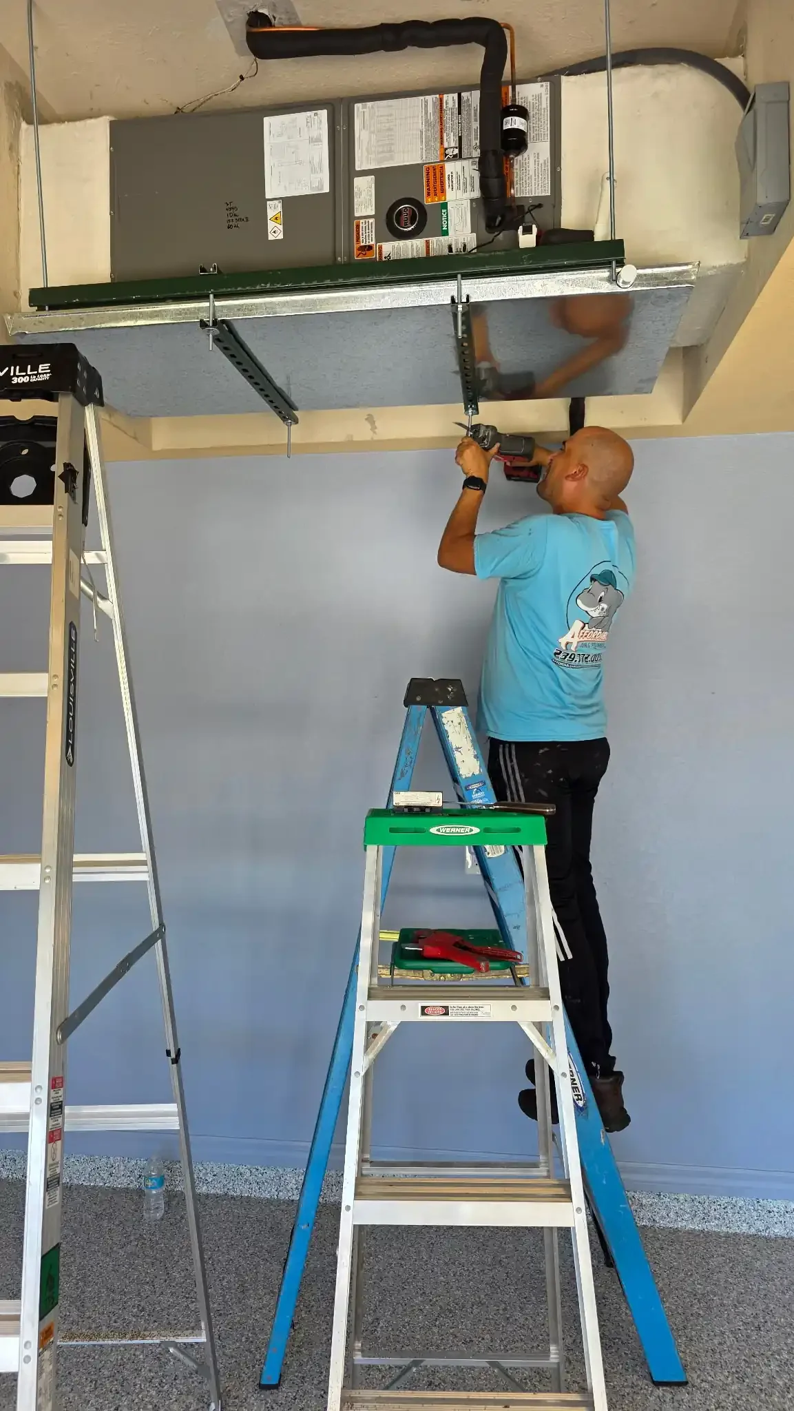 A professional technician from Affordable Air and Plumbing performs an AC installation in a garage. He is standing on a blue stepladder, working on a ceiling-suspended HVAC system with insulated refrigerant lines and a secondary drain pan.