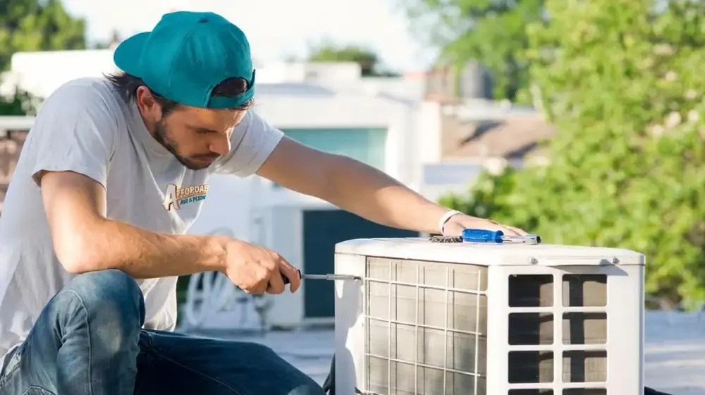 An Affordable Air and Plumbing Technician is working on unscrewing the faceplate of an outdoor heat pump unit in order to perform repairs. The technician wears a gray shirt and teal hat.