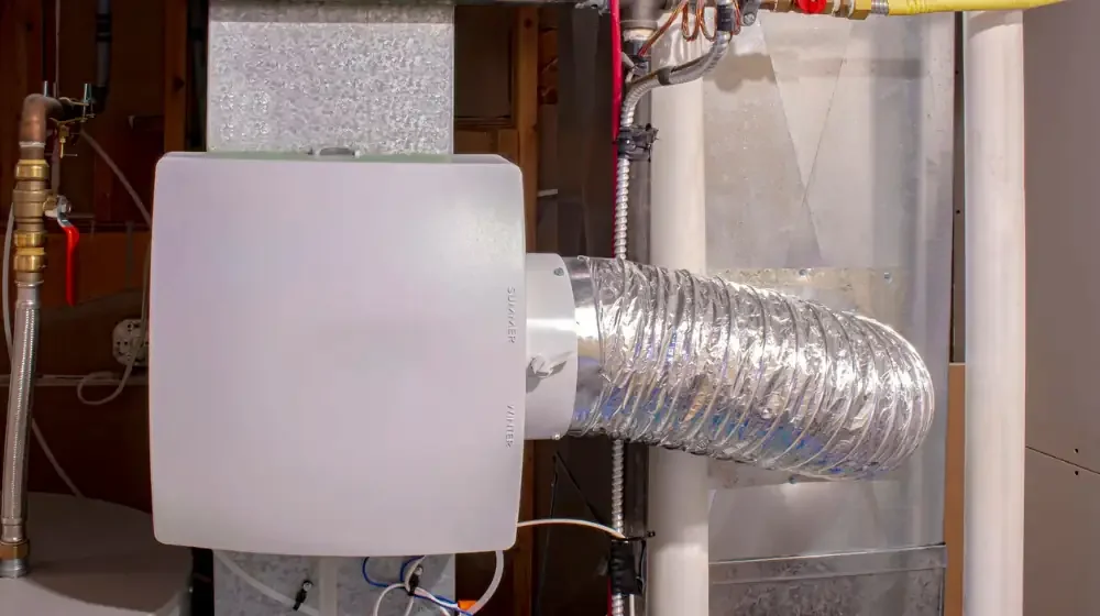 A gray whole home dehumidifier is attached to a central air conditioning system via tubing and wiring.