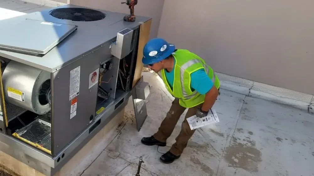 An Affordable Air and Plumbing Technician working on a commercial roof top unit Air Conditioner. He is preparing to perform a repair and has a few documents on the specific AC unit in his left hand.