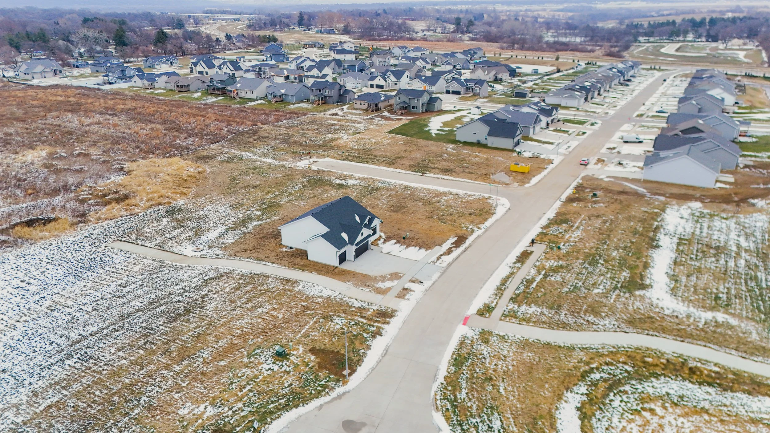 An aerial view of a residential neighborhood with many newly built houses, paved roads, and some snow on the ground. There are open areas of land with little to no development.