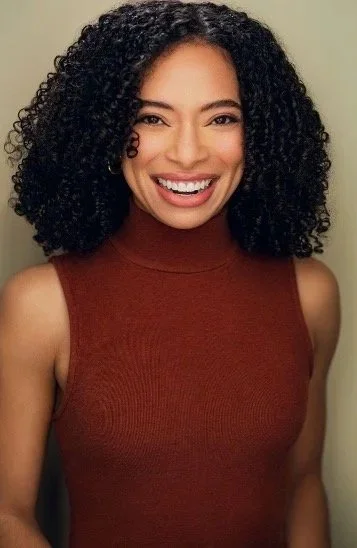 A woman with curly black hair smiling, wearing a sleeveless maroon top, standing indoors against a beige wall.