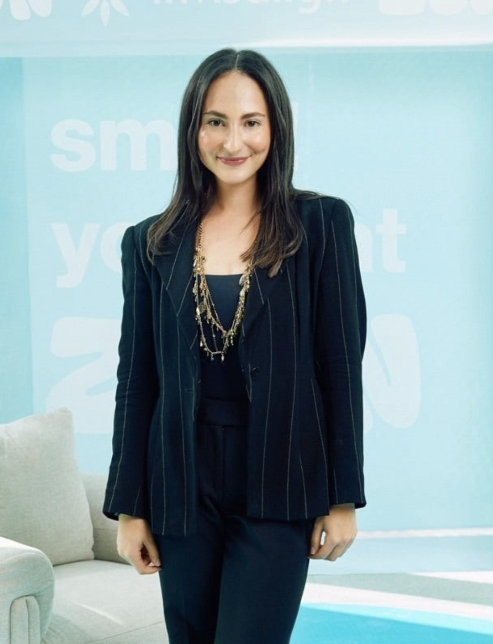 A woman with dark hair in a dark pinstripe blazer and matching pants, wearing a layered gold necklace, standing in a light blue room with a light-colored sofa in the background.
