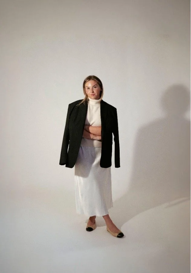A woman standing in front of a plain background, wearing a black blazer, white turtleneck, a long white skirt, and beige heels.