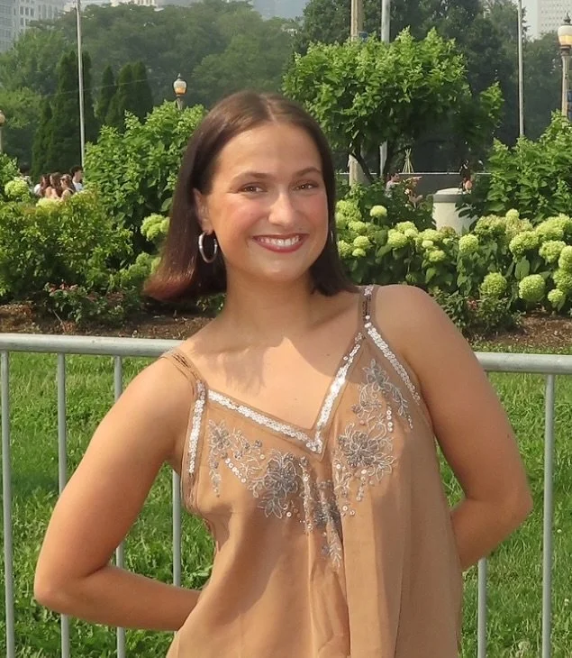 A young woman with shoulder-length brown hair smiling at the camera, wearing a beige spaghetti strap top with embroidery, standing outdoors in a park with green bushes, trees, and a metal fence behind her.