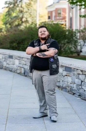 A person with a beard and long hair posing outdoors on a sidewalk, wearing a black shirt, gray pants, and a black vest, making a peace sign and smiling.