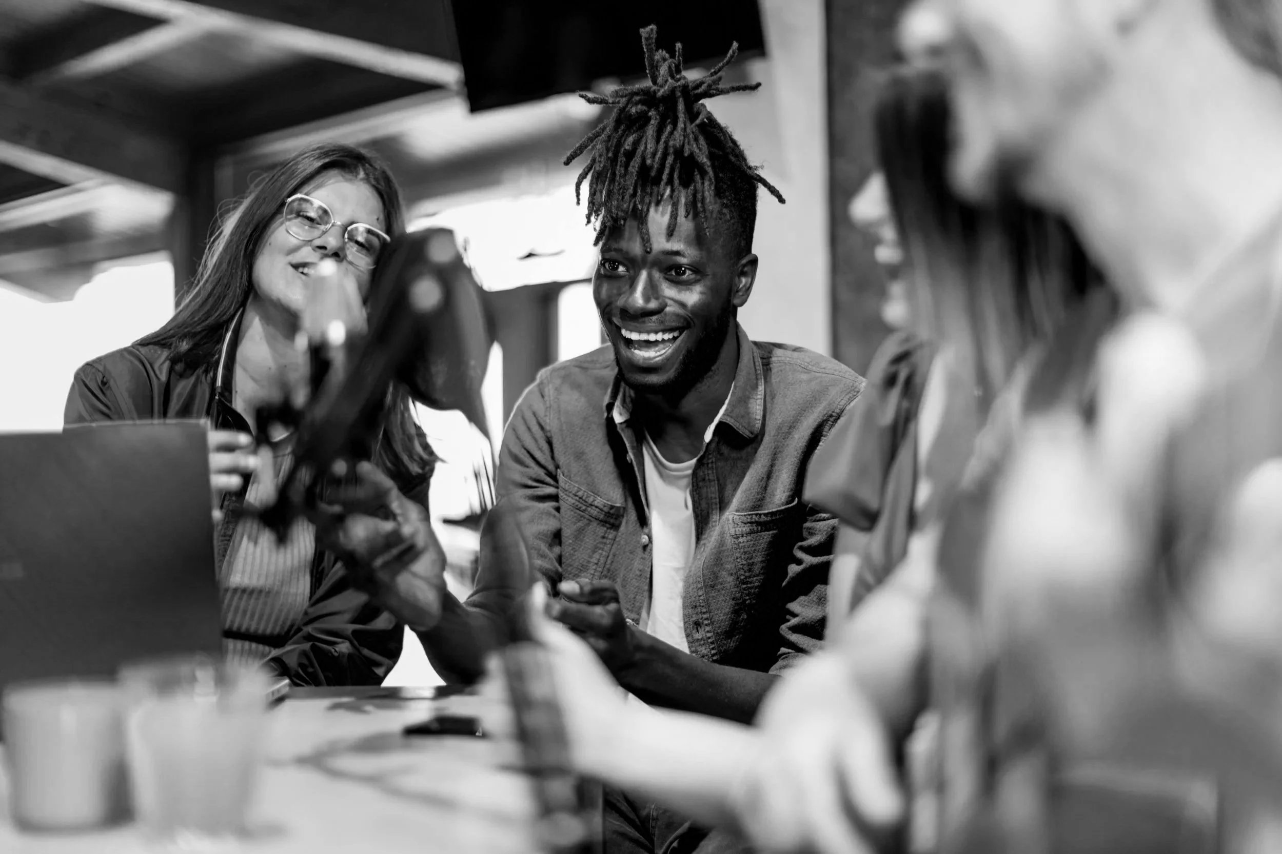 A group of diverse people sitting around a table, smiling and engaging in conversation, with one person showing something on a phone or tablet.