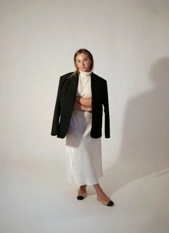 Woman standing against a plain light background, wearing a white dress, a black blazer draped over her shoulders, and black-and-white heels, with arms crossed.