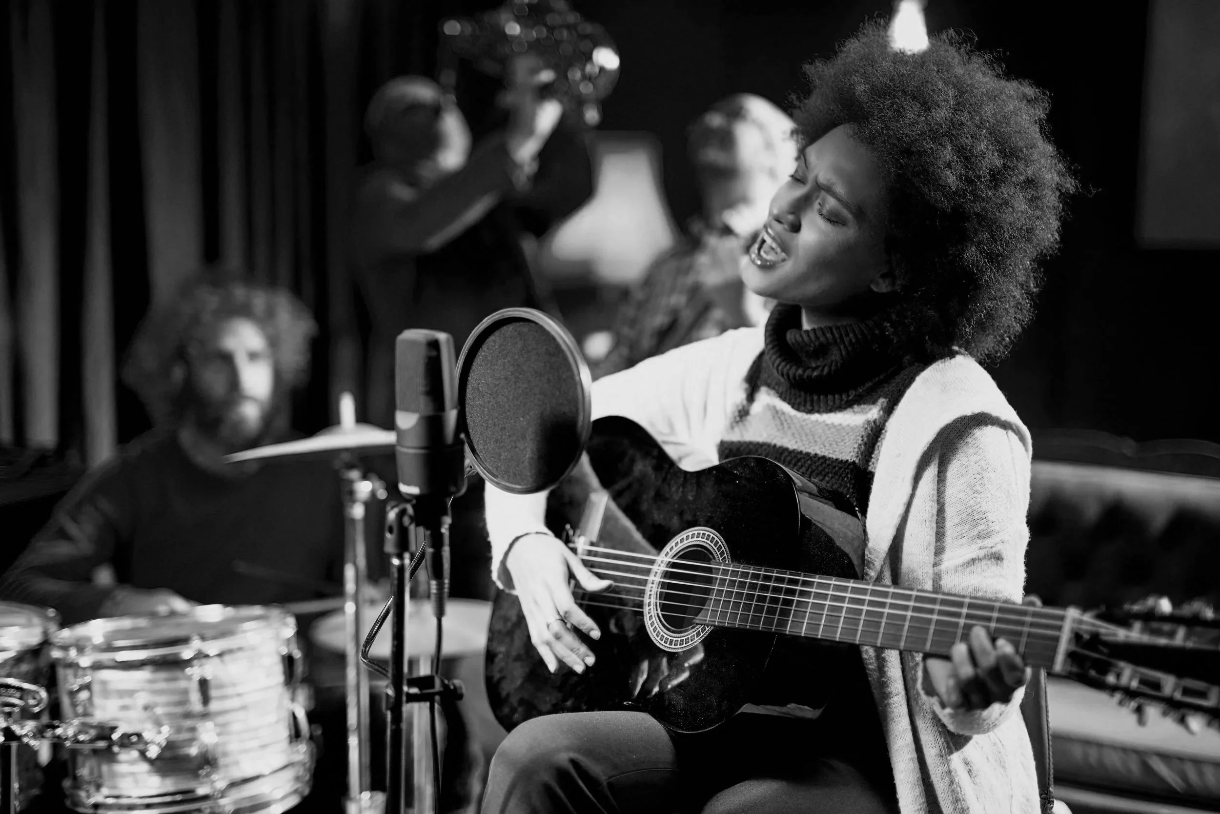 A woman singing and playing guitar in a recording studio with a microphone in front of her, while a drummer plays drums in the background.