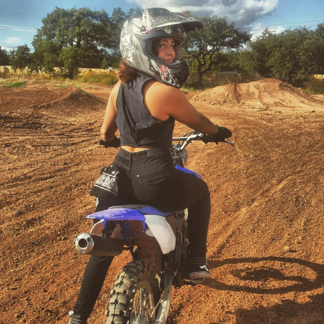 A woman wearing a helmet sits on a dirt bike, looking over her shoulder, with dirt mounds and trees in the background.