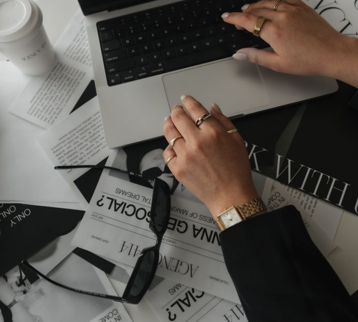 A person's hands with rings and a watch resting on a laptop keyboard, surrounded by reading glasses, papers, and magazines on a desk.