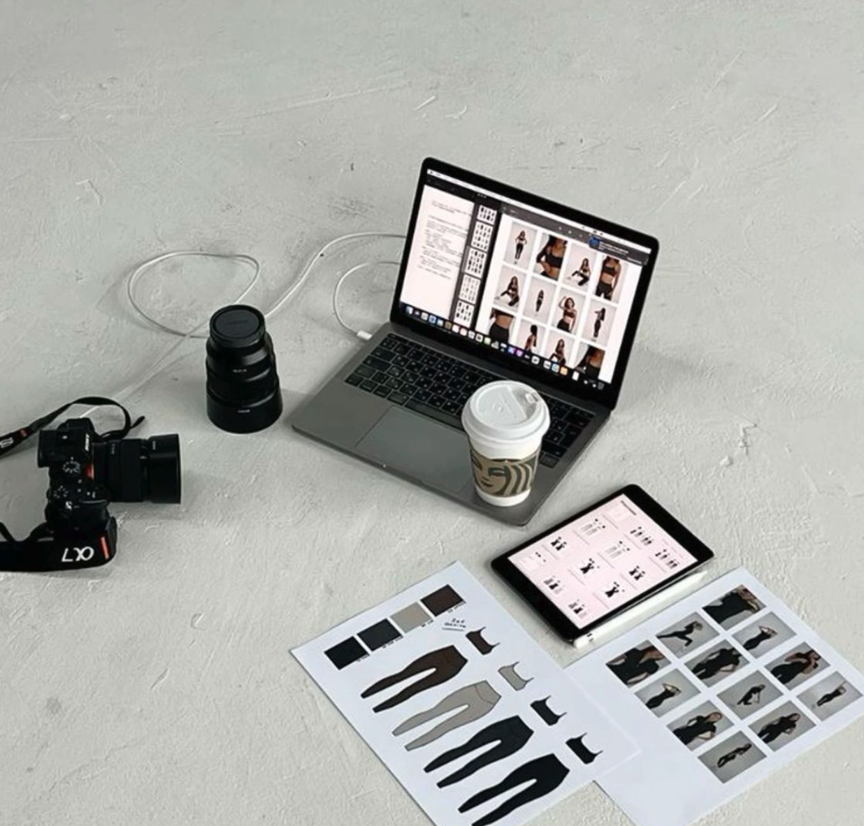 Photograph of a creative work setup showing a laptop displaying a fashion photo shoot, a tablet with editing interface, a printed mood board with clothing sketches and images, a camera, a camera lens, and a cup of coffee on a white textured surface.