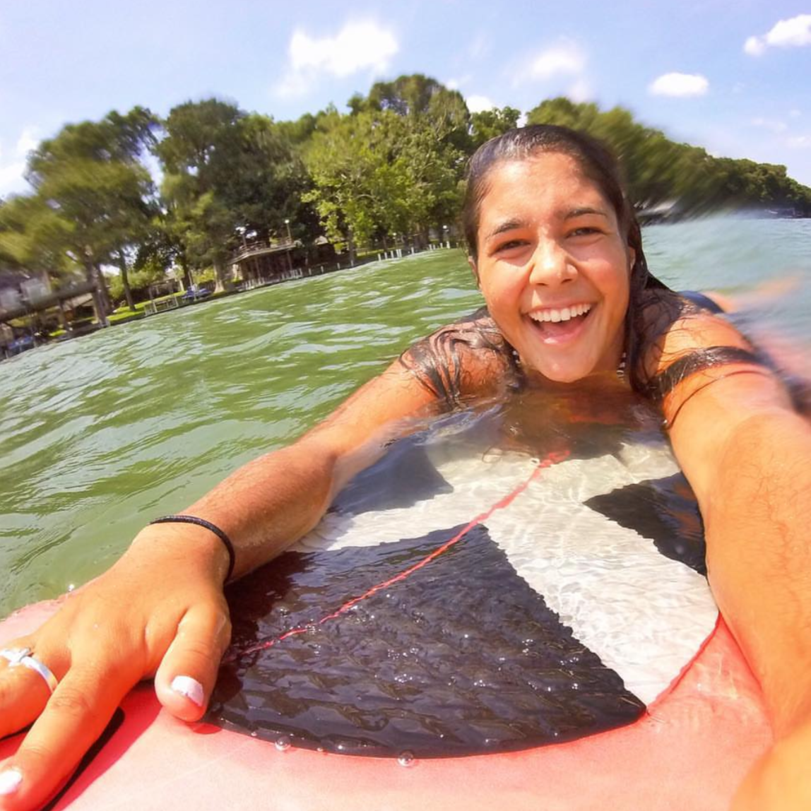 Woman smiling and taking a selfie while lying on a paddleboard in a lake, surrounded by trees and blue sky.
