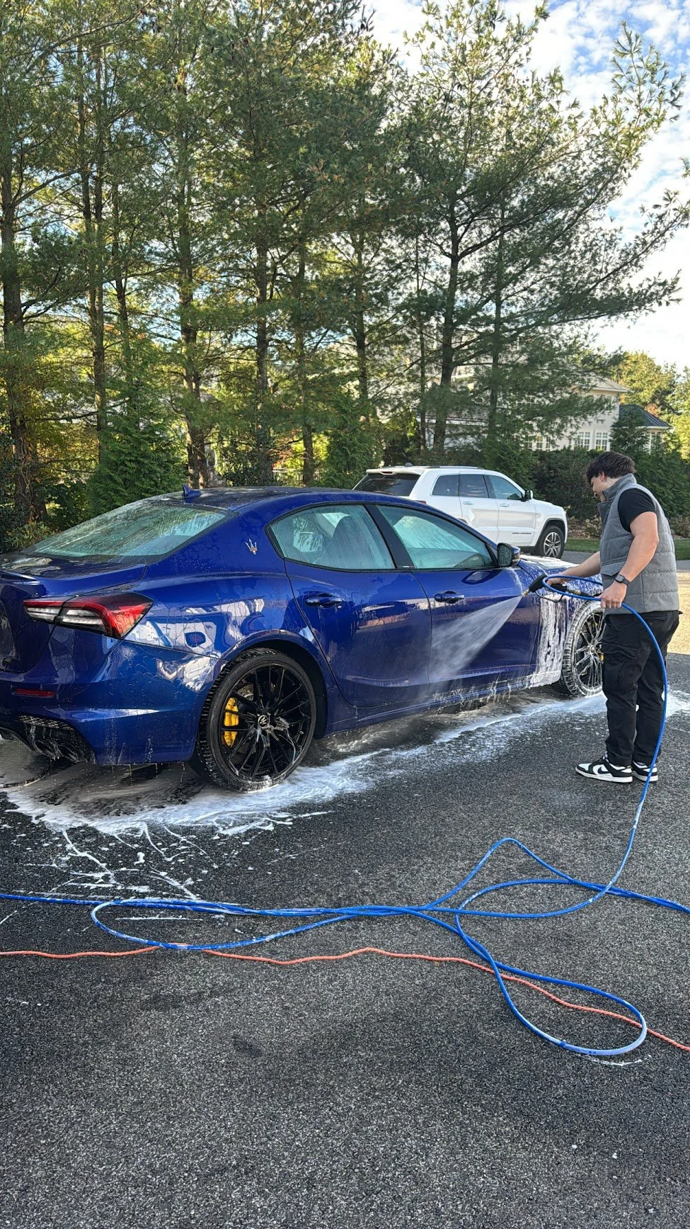 A expert detailing a blue Maserati sports car professionally in a driveway, with trees and a house in the background.