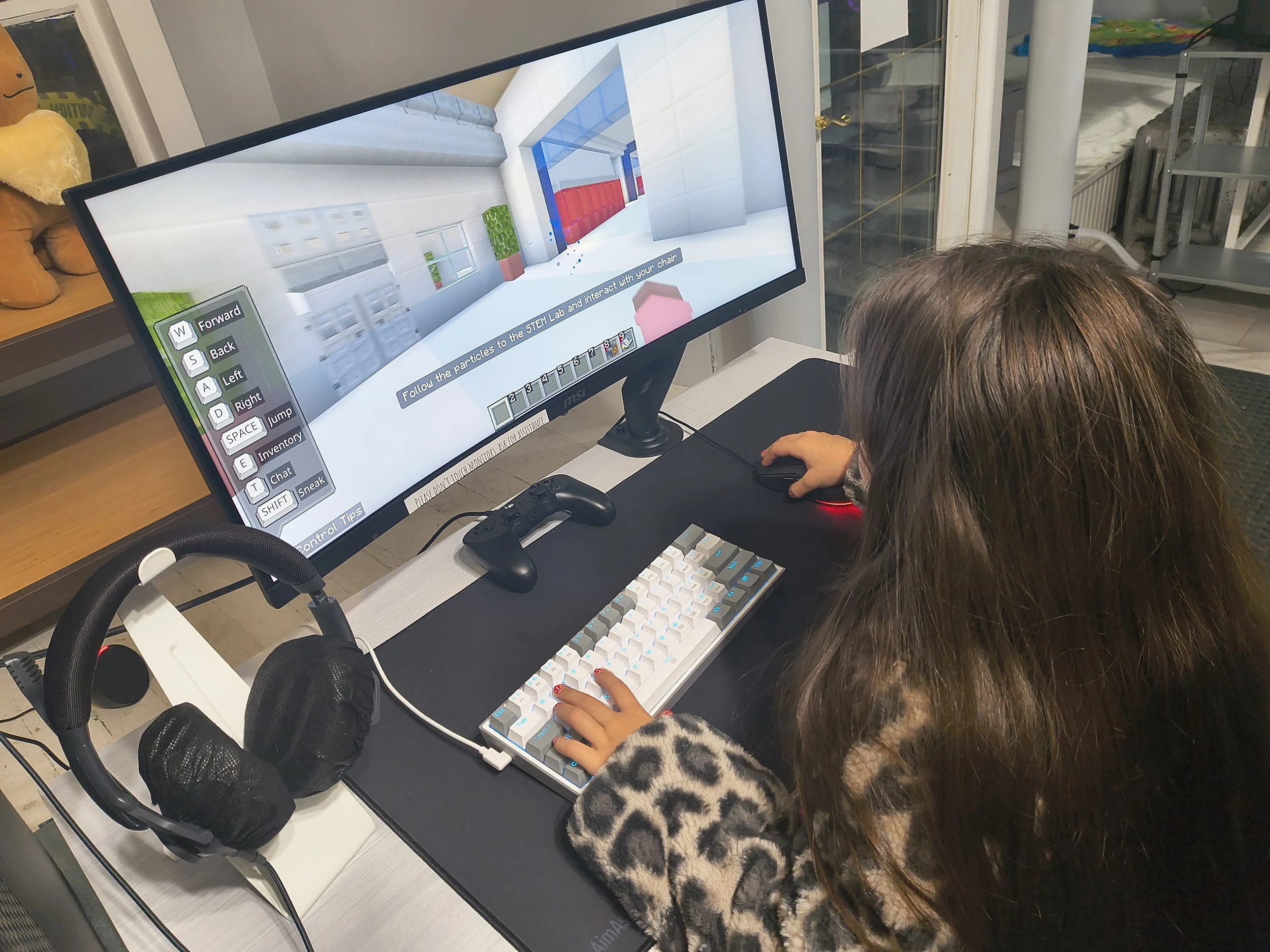 A young girl is playing a computer game at a desk with a keyboard, mouse, and headphones, in a room with shelves and a window in the background.