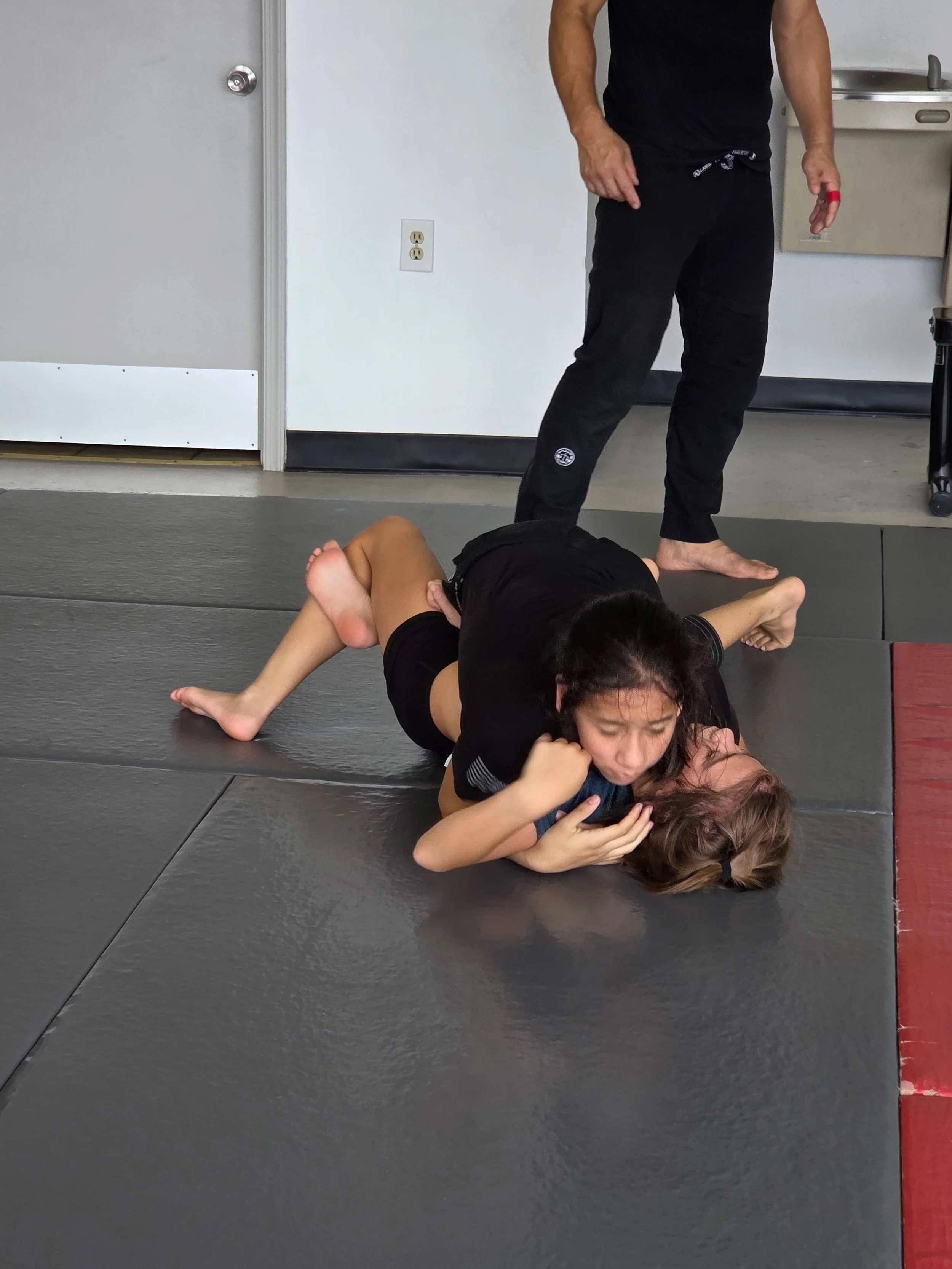 Two children practicing Brazilian Jiu-Jitsu on mats while a coach observes.