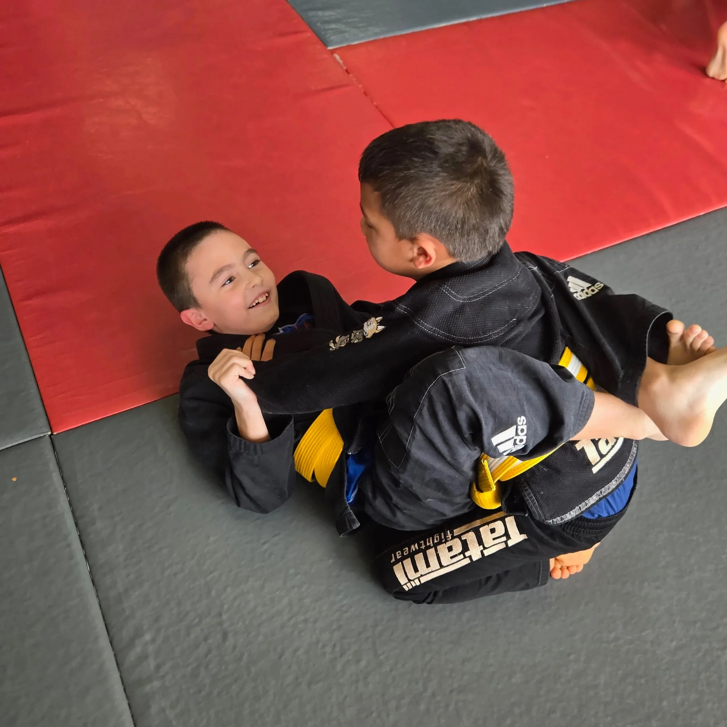 Two young boys practicing Brazilian Jiu-Jitsu on a padded mat, wearing gis with yellow belts, engaged in a playful grappling match.
