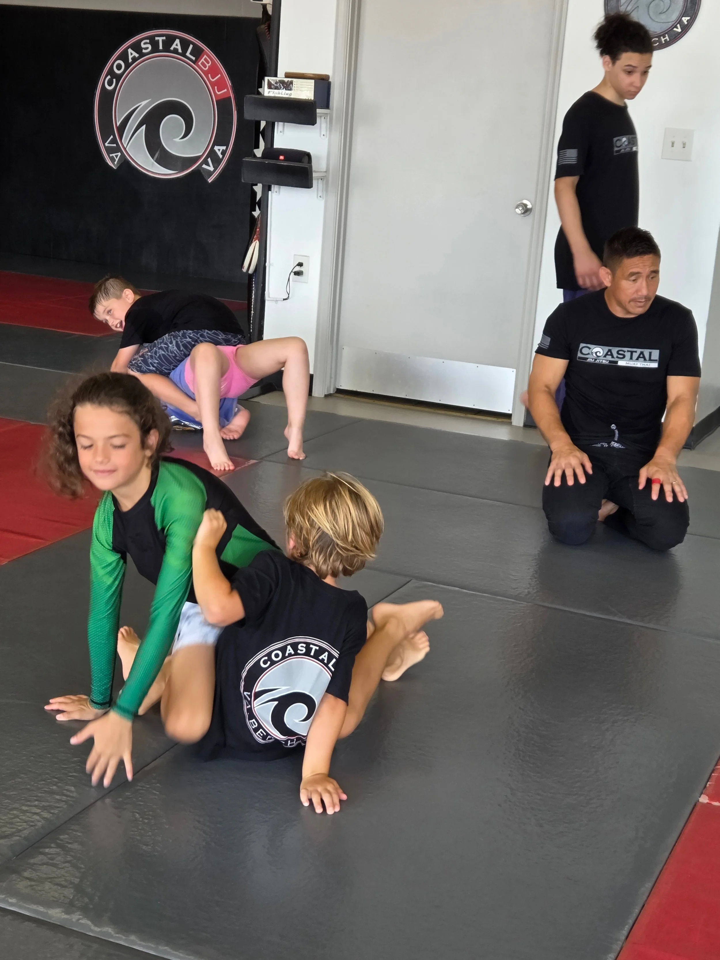 Kids practicing Brazilian Jiu-Jitsu on mats with instructor in a martial arts gym.