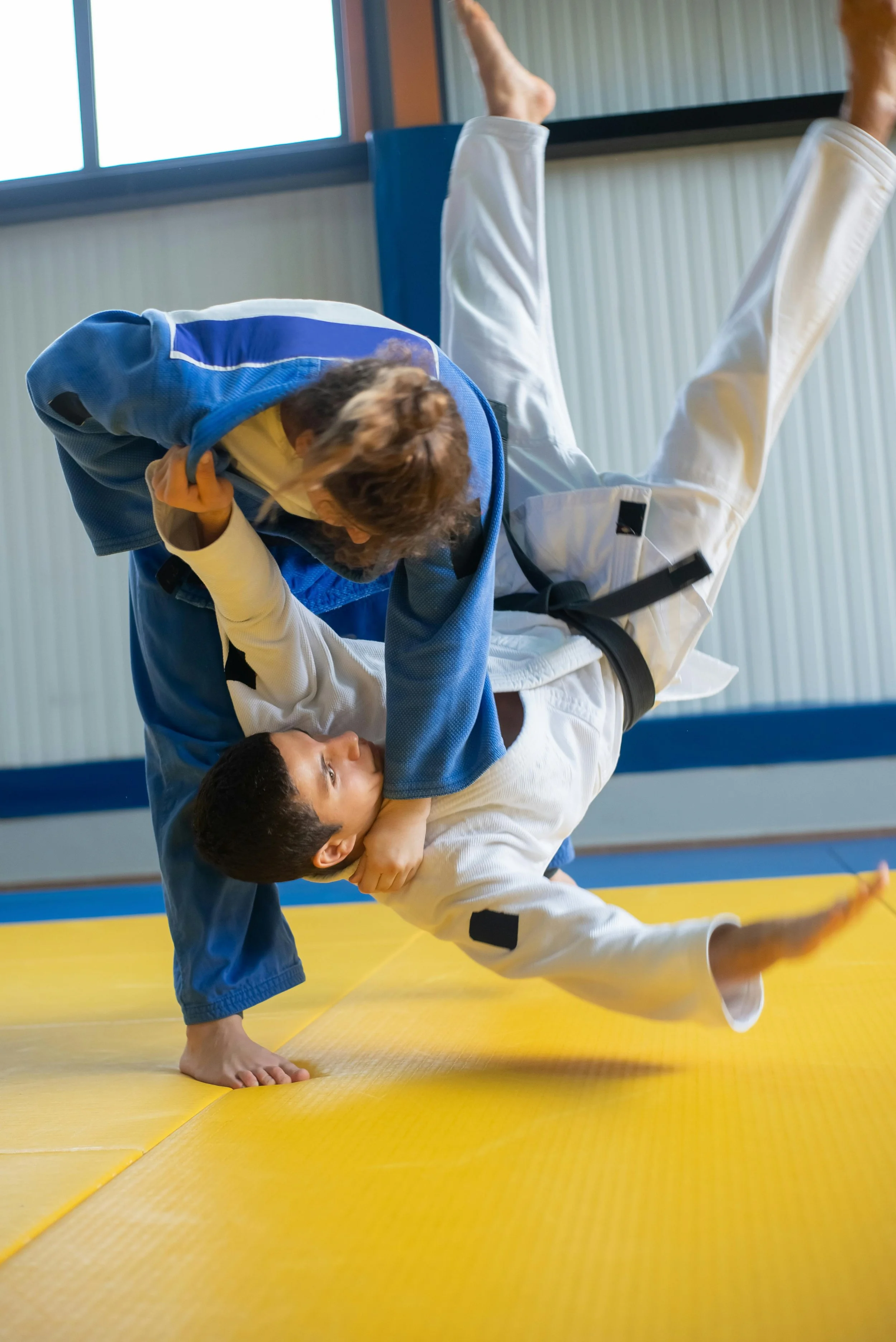 Two boys practicing judo on yellow mat, one in white gi and the other in blue gi, during a training session in gym.